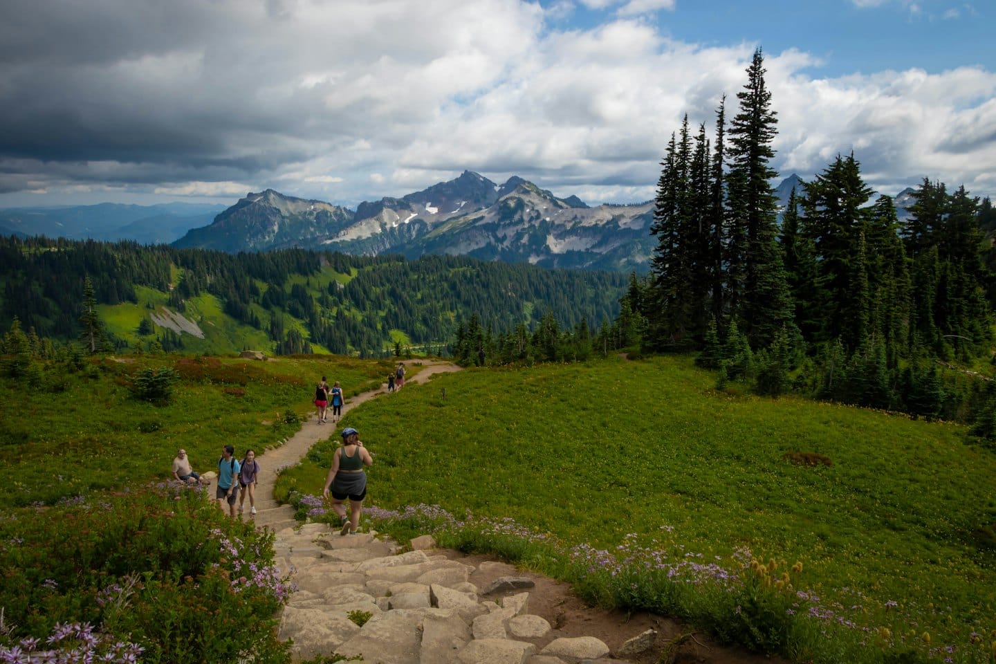 Hikers walk along a mountain path toward snowy peaks.