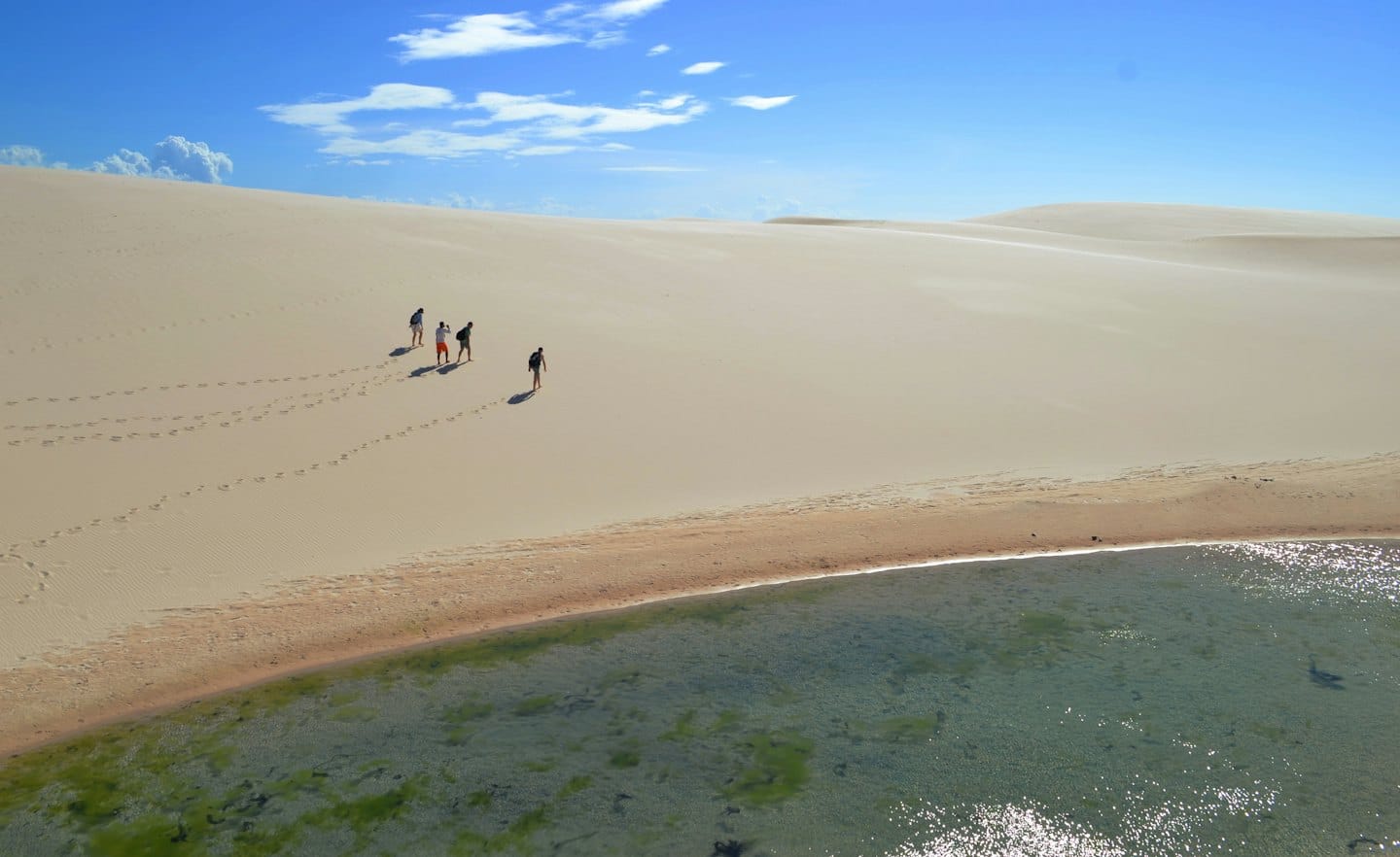 Four people walk across sand dunes beside a shallow lagoon.